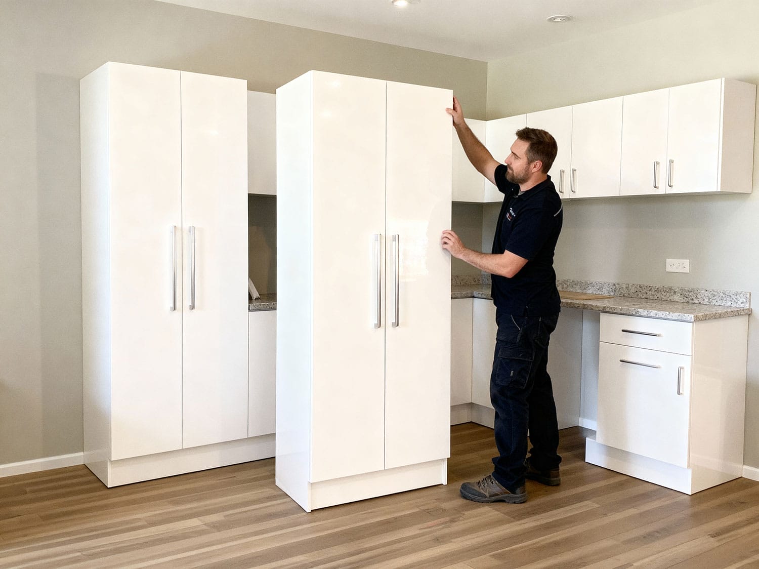 Installer positioning a tall pre-assembled white gloss kitchen cabinet in a modern showroom with matching wall and base units