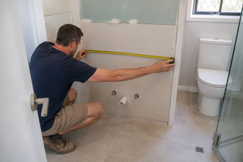 Homeowner measuring bathroom vanity space with plumbing, doorway and shower clearance visible