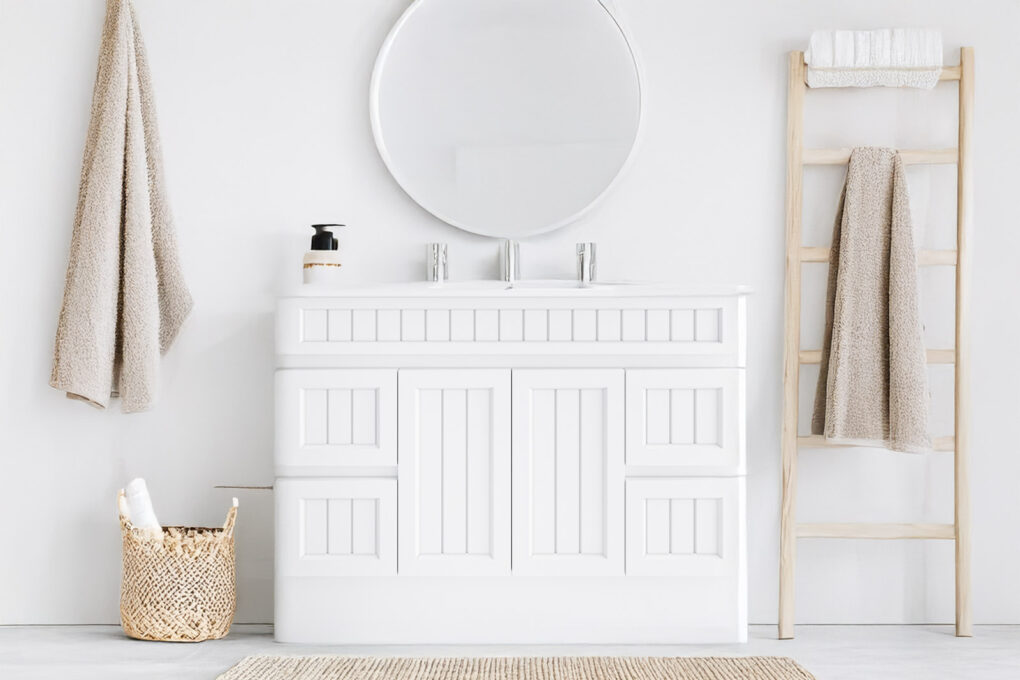 White Hamptons-style single vanity unit with a round mirror, set in a bright bathroom with neutral decor and natural textures.