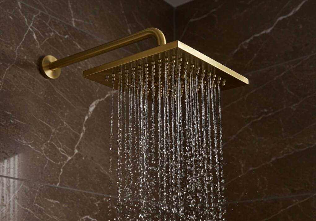 Gold square rain shower head with water flowing, mounted on a brown marble bathroom wall