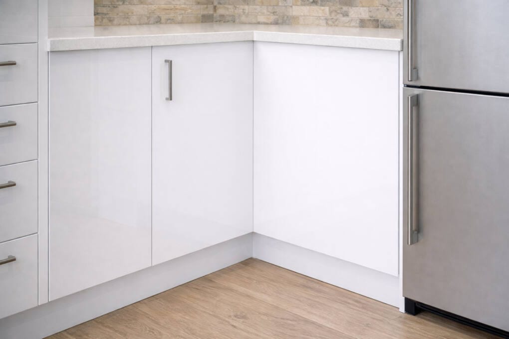 White gloss kitchen corner cabinet with integrated handle, stone benchtop above, and adjacent stainless steel refrigerator.