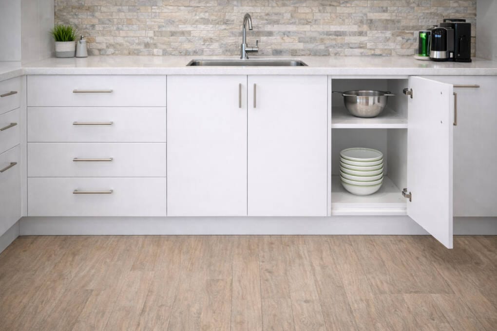 White kitchen base cabinets with drawers and cupboard storage beneath a sink, featuring a stone benchtop, tiled splashback, and open cabinet displaying cookware and bowls.