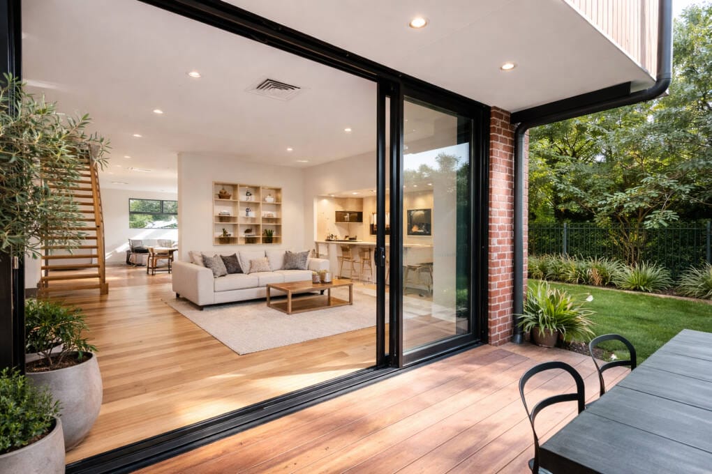 Black aluminium stacking doors fully open to an Australian open-plan living area with timber floors, modern lounge, and seamless indoor–outdoor connection to a garden and deck