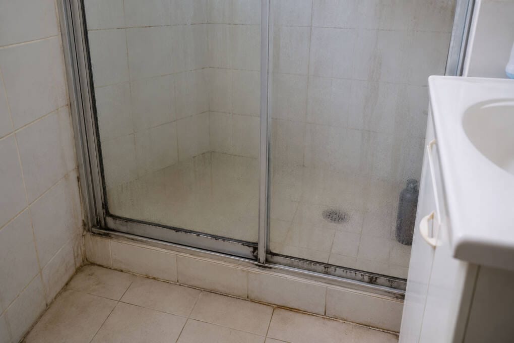 Older framed shower screen with foggy glass, worn seals and stained shower track before replacement